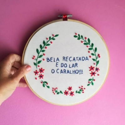 Aro de madeira com bordado floral e texto em português contra parede rosa