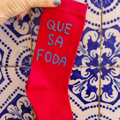 Meia vermelha com bordado azul 'QUE SA FODA' segurada à mão sobre azulejos azuis e brancos