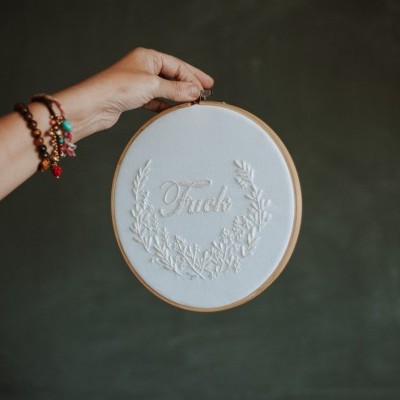 Bastidor de madeira com tecido branco bordado com a palavra 'Fuck' e folhas, segurado por mão com pulseiras.