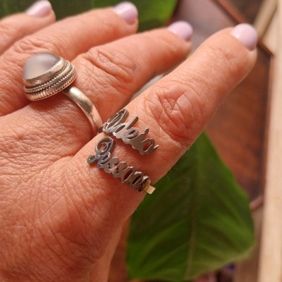 Mão segurando anéis de prata, um com pedra opaca e outro com palavras Vida e Pássaro, planta ao fundo