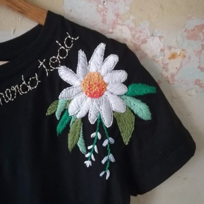 T-shirt preta com bordado de flor branca e folhas verdes no ombro e texto bordado branco na gola