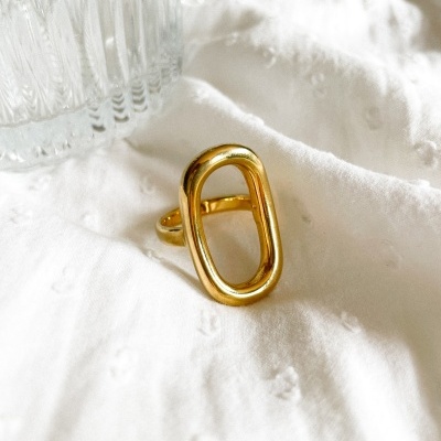 Anel dourado oval sobre tecido branco com vidro transparente ao lado.