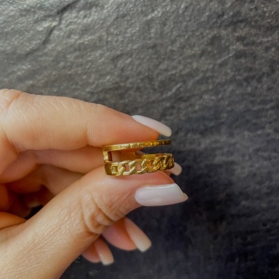 Anel dourado com design de corrente sendo segurado por uma mão com unhas brancas.
