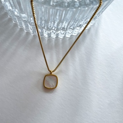 Colar dourado com pendente quadrado translúcido sobre superfície branca e objeto de vidro