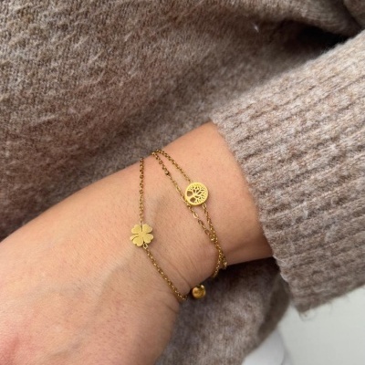 Mão com pulseiras douradas finas com pendentes de trevo e árvore da vida, anel dourado, camisola bege e calças brancas