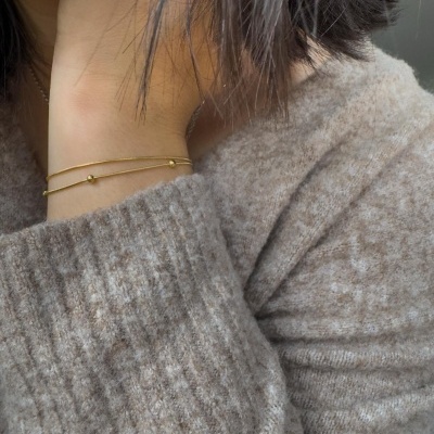 Pulseira fina dourada com pequenos enfeites esféricos no pulso de pessoa com camisola cinza de malha
