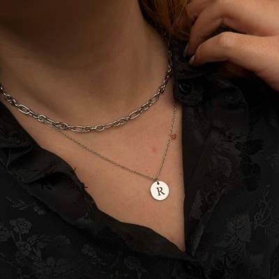 Colar em metal prateado com pendente redondo gravado R e colar de elos maiores em pescoço com camisa preta floral.