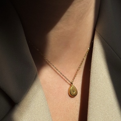 Colar dourado com pendente em forma de gota no pescoço de uma pessoa com casaco bege.