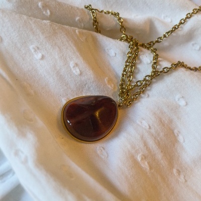 Colar dourado com pendente de pedra castanha sobre tecido branco texturado