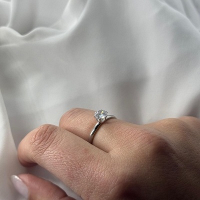 Anel solitário prateado com pedra transparente num dedo com unhas nude sobre tecido branco