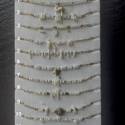 Doze pulseiras brancas com contas e elementos dourados em suporte bege de tecido linho.