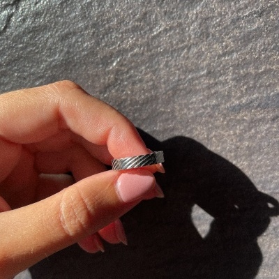 Anel prateado com padrão de riscas diagonais segurado por uma mão com unhas pintadas de rosa claro