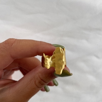 Anel dourado em metal com design irregular segurado por mão com unhas verdes em fundo branco.
