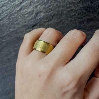 Anel dourado largo com textura granulada no centro no dedo médio de uma mão