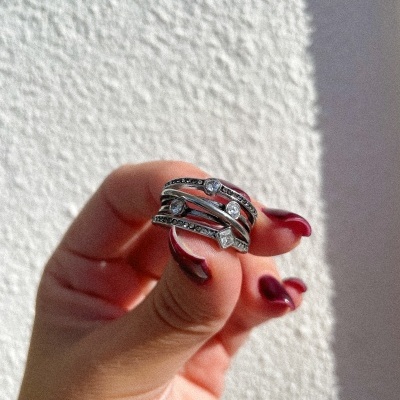 Anel prateado com várias fitas e brilhantes segurado por mão com unhas pintadas