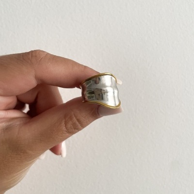 Anel largo prateado com bordas douradas segurado por uma mão em fundo branco