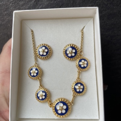 Conjunto de colar e brincos dourados com motivo floral azul e branco, em caixa branca.