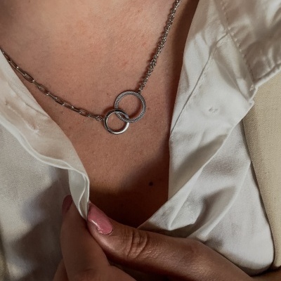 Colar com dois pingentes de argolas entrelaçadas prateadas no pescoço de uma pessoa com camisa branca.