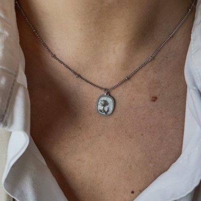 Colar prateado com medalha de rosa em relevo no pescoço de pessoa com camisa branca.