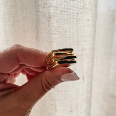 Anel dourado moderno com três bandas brilhantes nas mãos à frente de cortina clara