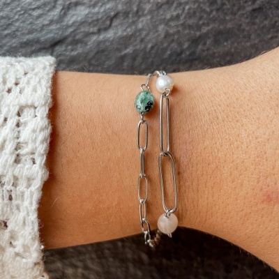 Braceletes de metal prateado com pedras verdes, pérola e pedra rosa no pulso com manga de malha branca