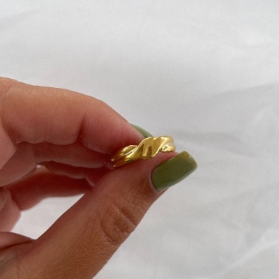 Anel dourado ondulado segurado por mão com unhas verdes, fundo branco liso