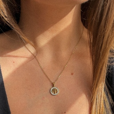 Colar dourado com pendente redondo de árvore, usado por pessoa com cabelo louro e camisa preta