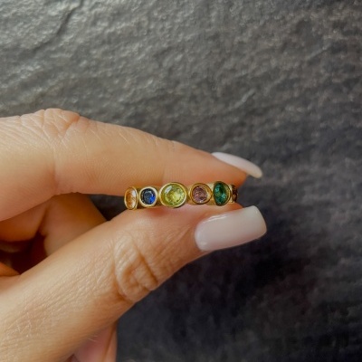 Anel dourado com pedras coloridas segurado por uma mão com unhas compridas brancas