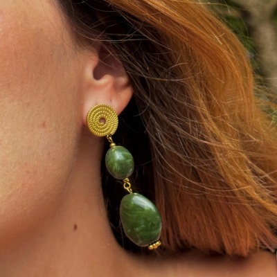 Brinco dourado com pedras verdes pendurado na orelha de pessoa com cabelo ruivo