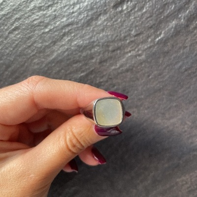 Anel com pedra quadrada esbranquiçada segurado por mão com unhas vinho