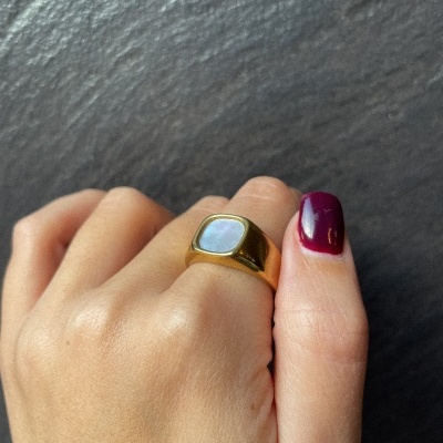 Anel dourado com pedra quadrada opalescente numa mão com unha bordô