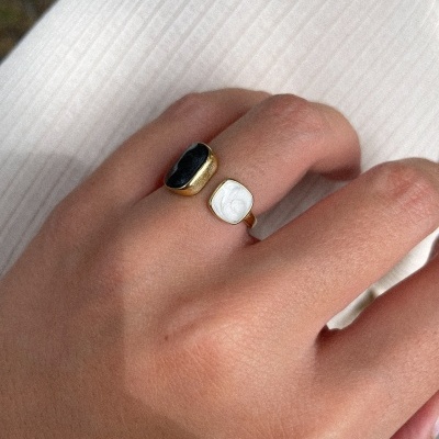 Anel dourado com duas pedras preta e branca em mão com fundo branco canelado