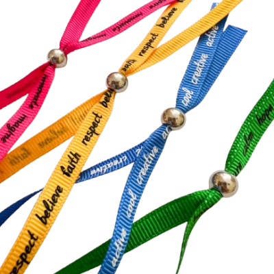 Pulseiras coloridas de tecido com palavras motivacionais em preto e bolas metálicas como fechos.