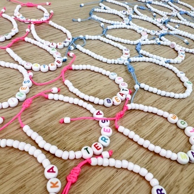 Pulseiras de contas brancas com letras coloridas e cordões rosa e azul sobre mesa de madeira