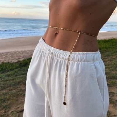 Cinto dourado de corrente fina com pendente flor, cintura com calças brancas, praia ao fundo
