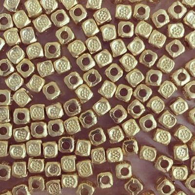 Contas quadradas douradas com padrão saliente e furo central em superfície rosa