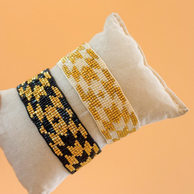Duas pulseiras de contas com padrão xadrez, dourado e preto e dourado e branco, numa almofada cinza.
