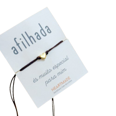 Pulseira com cordão preto e pendente dourado em forma de coração num cartão branco com texto