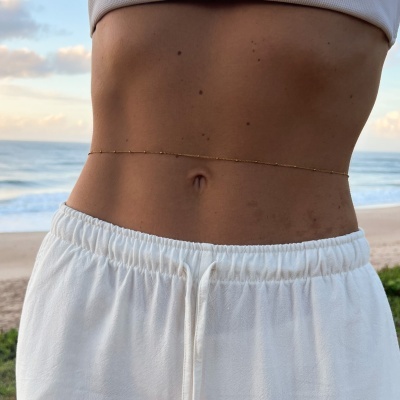 Corrente dourada na cintura de pessoa com roupa branca na praia ao pôr do sol
