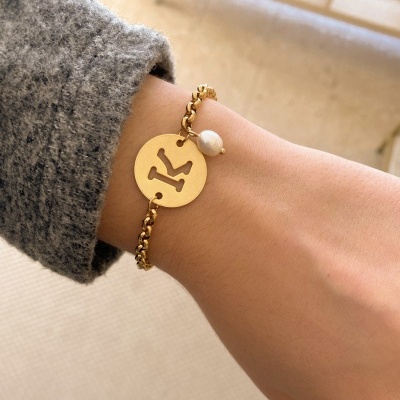 Pulseira dourada com letra K e detalhe de pérola no pulso.