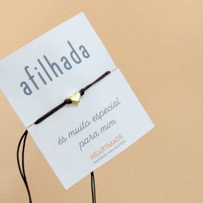 Pulseira com cordão castanho escuro e peça dourada em forma de coração com cartão branco