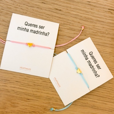 Pulseiras rosa e azul clara com coração dourado em cartões com texto Queres ser minha madrinha? sobre mesa de madeira.