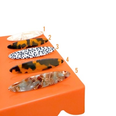 Cinco presilhas de cabelo ovais em acrílico com diferentes padrões de cor sobre suporte laranja.