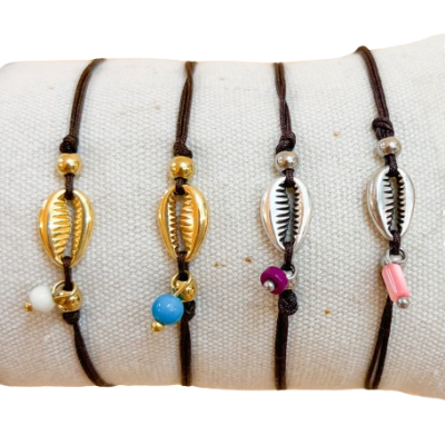 Pulseiras com cordão castanho e conchas metálicas douradas e prateadas com contas coloridas sobre tecido bege.