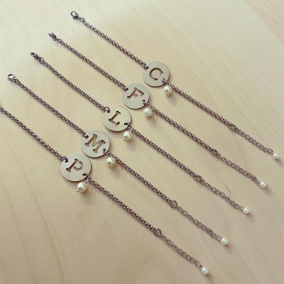 Cinco pulseiras metálicas com discos com letras e pérolas sobre fundo de madeira.