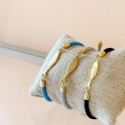 Três pulseiras com corda azul, cinza e preta com peixes dourados, sobre almofada bege trançada.
