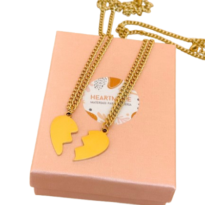 Dupla de colares dourados com pendentes amarelos de coração partido sobre caixa rosa com etiqueta HEARTMADE.