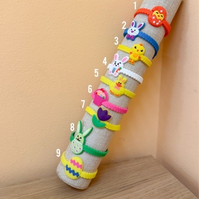 Dez pulseiras coloridas com motivos de Páscoa expostas num suporte de tecido cinzento.