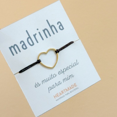 Pulseira com cordão preto e coração dourado num cartão branco com texto
