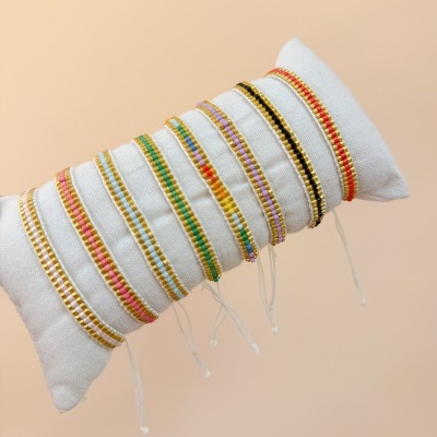 Pulseiras coloridas com fios e detalhes dourados exibidas num apoio de tecido branco.